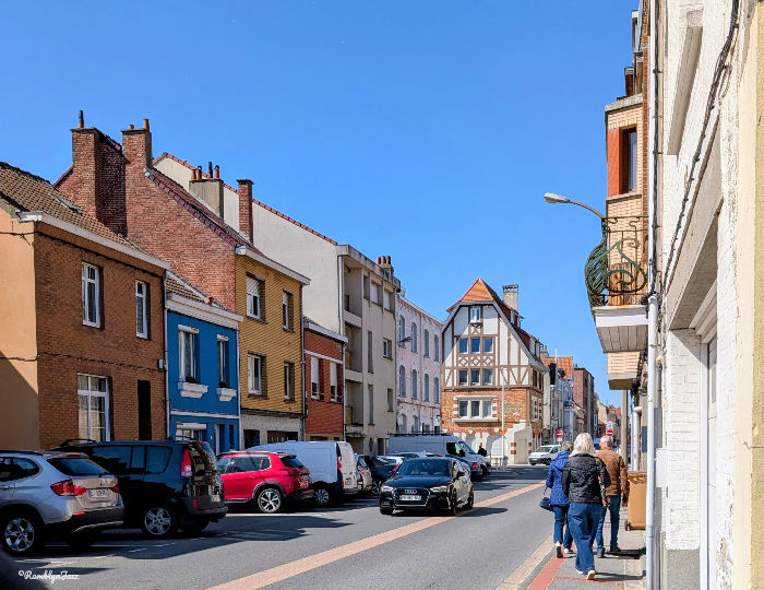 Sunny street with colourful row houses, parked cars, and two people walking on the sidewalk under a clear blue sky.