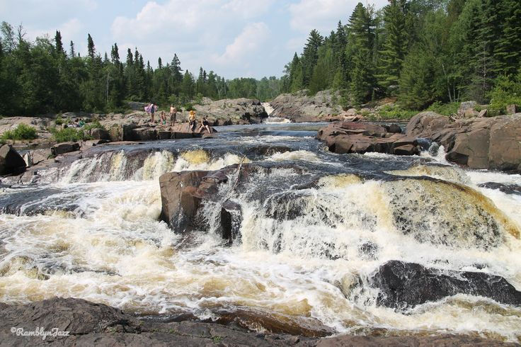 Rugged waterfall with clear rushing water, surrounded by rocks and lush green forest. People relax and swim near the falls under a bright sky.