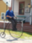 Man riding a penny-farthing bicycle in a historical setting. He's wearing a blue shirt. Sign reads "MEALS." Sunny day, vintage vibe.