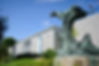 Bronze cougar sculpture poised on a rock, set against a modern, white brick building with lush greenery and a clear blue sky in the background.