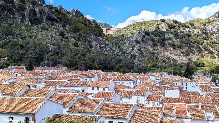 White houses with red-tiled roofs nestled in a valley. Green rocky hills visible under a blue sky with clouds. Calm and picturesque setting.