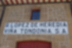 The entrance to a winery. The wall is made of different sizes and shapes of sand-coloured brick and white grout. A large white sign with black block letter printing reads "R. Lopez de Heredia Vina Tondonia. S.A."