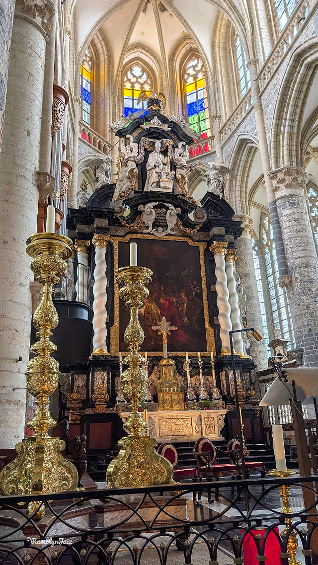 Ornate cathedral altar with gilded candlesticks, crucifix and painting beneath colorful stained-glass windows in a solemn nave.
