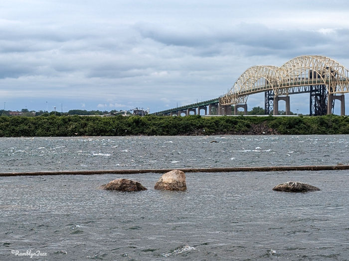 Cloudy sky over a river with partially submerged rocks in the foreground. An arched bridge spans the background. Calm, overcast mood.