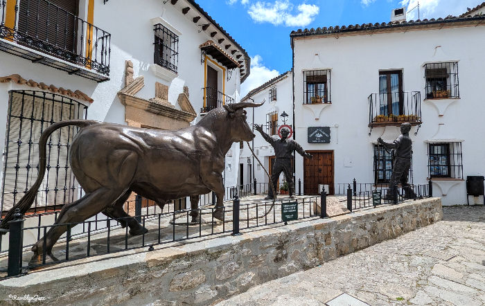 Bronze bull statue and two matador statues in a white courtyard, surrounded by traditional Spanish buildings, under a blue sky.