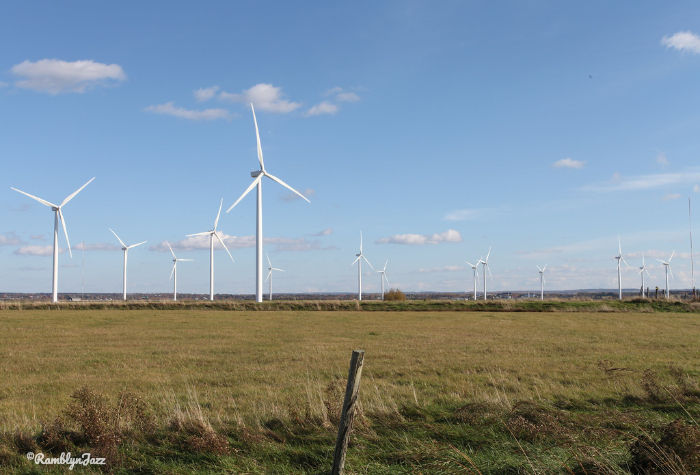 Wind turbines in a green field under a clear blue sky with scattered clouds. A fence post stands in the foreground.
