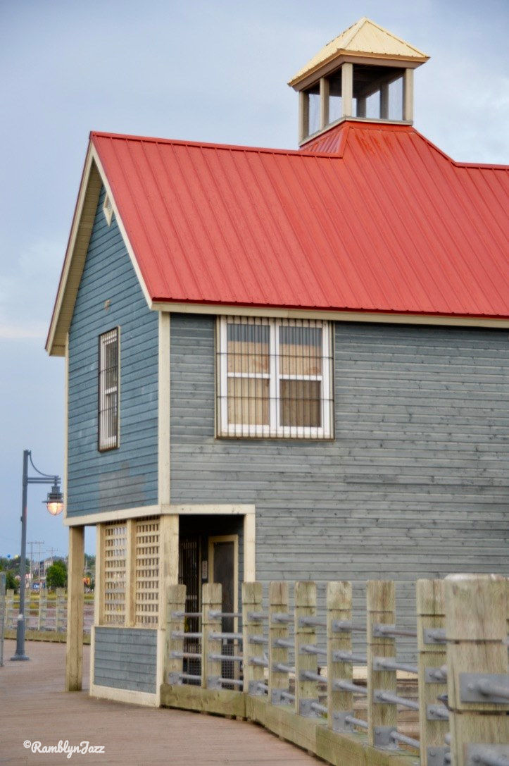 Blue building with a red roof on a boardwalk, under a cloudy sky. A vintage lamp post is visible. Calm and serene atmosphere.