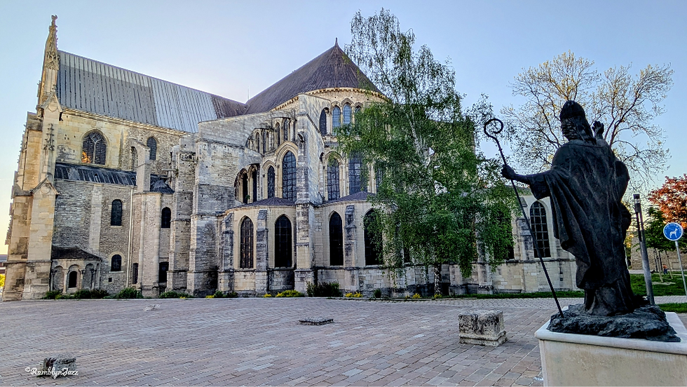 Historic stone cathedral courtyard with a hooded bishop statue holding a staff, framed by trees under a clear blue sky.