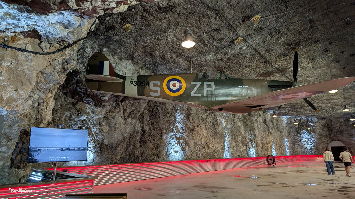 A vintage military aircraft is displayed in a rocky cave setting, lit by overhead lights. Red and white patterns are on the floor.