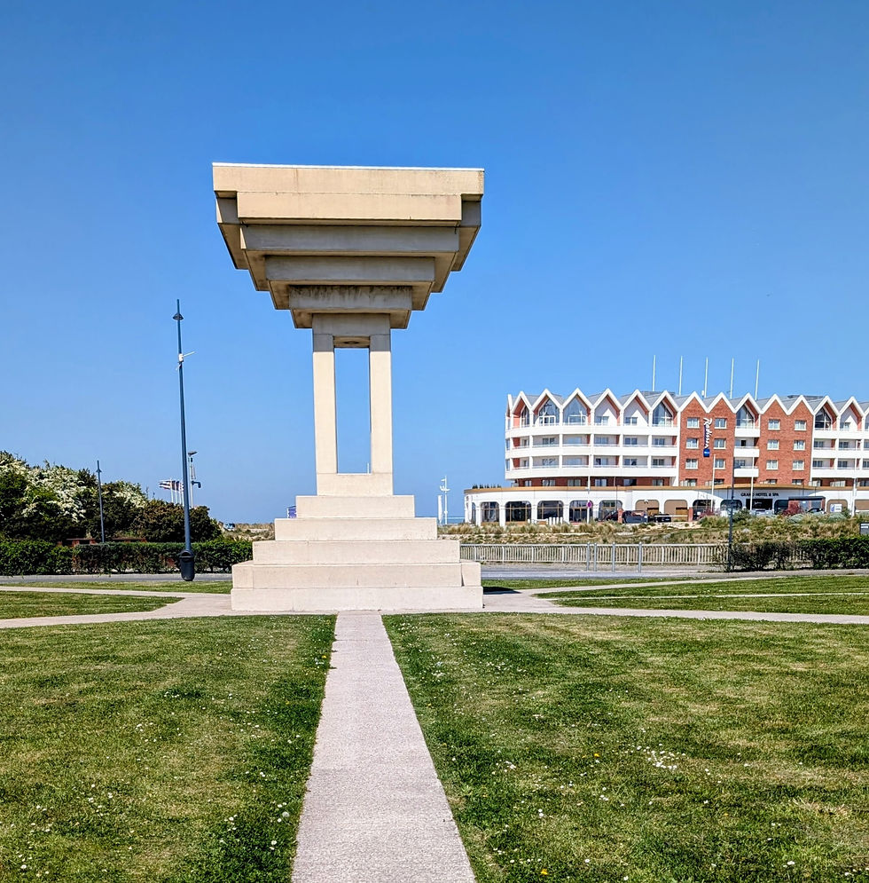 Abstract concrete monument on a grassy seaside lawn, with a hotel and clear blue sky in the background.