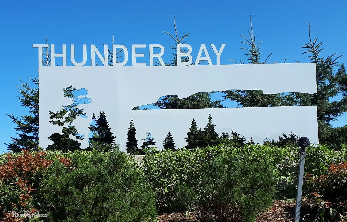 Sign reading "THUNDER BAY" with tree silhouettes, set against a clear blue sky and green foliage, evokes a serene and natural mood.