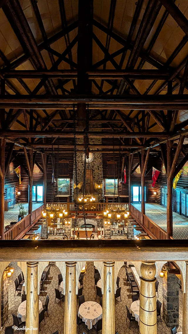 Rustic lodge interior with wooden beams, chandeliers, and stone walls. Round tables with white cloths, flags, and paintings visible. Cozy ambiance.