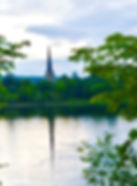 A church steeple reflected in a tranquil river, surrounded by lush green trees. The sky is cloudy, creating a peaceful, serene atmosphere.