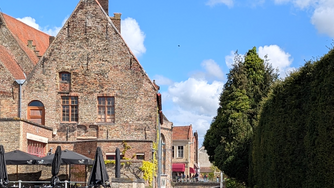Historic brick buildings by a canal under a blue sky. Black umbrellas cover a riverside patio. Verdant trees line the scene.
