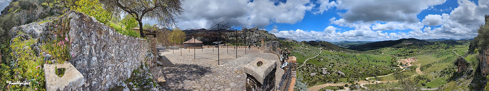 Panoramic view of ruins on a hilltop with stone walls, wildflowers, and people exploring. Vast green landscape under a blue, cloudy sky.