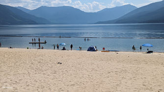 Sandy beach with people swimming and paddleboarding in a calm lake. Mountains and clouds in the background; blue tents and umbrellas dot the shore.