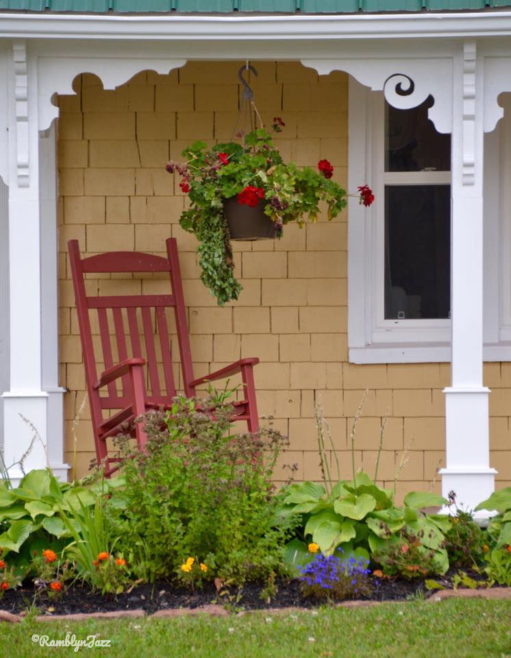 Red rocking chair and hanging flowers on a porch. Yellow shingles and lush greenery create a peaceful, sunny atmosphere.