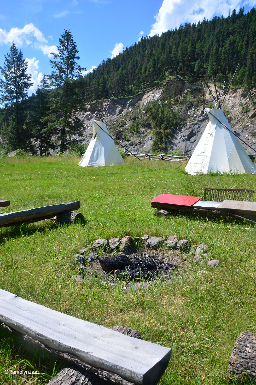 Two white tipis on grassy field with campfire pit and wooden benches. Forested hills in background under a bright blue sky.
