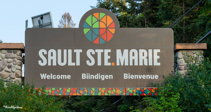 Sign reading "Sault Ste. Marie Welcome Biindigen Bienvenue" with colorful pattern, set in a lush green environment with trees.