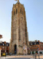 Tall historic brick clock tower in a quiet European square, with flags, nearby buildings, and a clear blue sky.