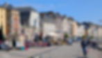 Sunny waterfront promenade with historic buildings, café seating, and pedestrians along a harbor under a clear blue sky.