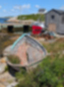 Weathered boat with peeling paint sits on grassy shore by the ocean. Red fishing boat and rustic buildings in the background under a blue sky.