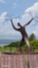 Metal sculpture of a tall figure with tree-like arms and smaller figures at the base, set outdoors with a scenic lake view and name plaques.