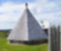 A wooden pyramid-shaped structure with a shingle roof stands in a grassy area. A picket fence and cloudy sky are in the background.