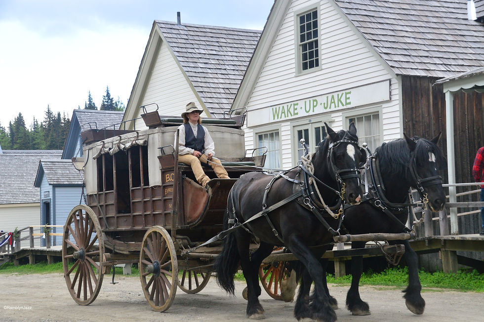 A stagecoach with driver pulled by two horses on a mud road in front of a line of 1800s wooden buildings