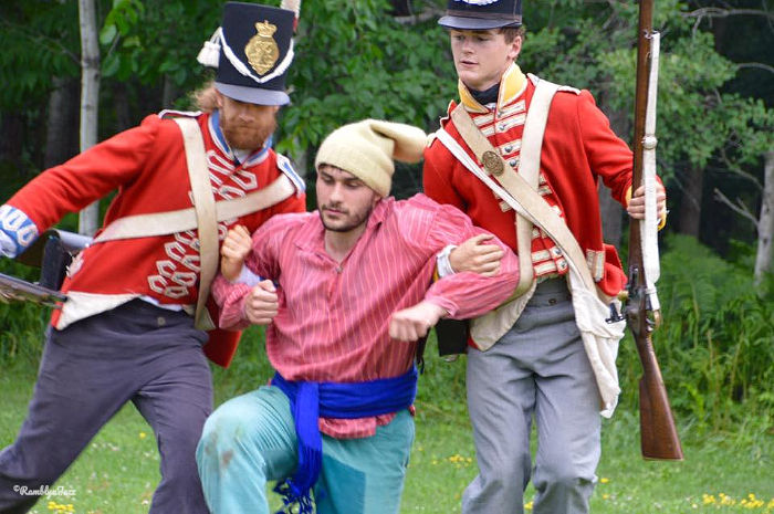 Two soldiers in red uniforms support a man in a pink shirt and beige hat. They stand on grass with trees in the background, appearing serious.