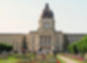 Large stone building with a dome, flanked by flags, stands amidst colourful flower gardens and pathways on a sunny day.