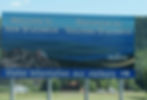 Billboard reads "Welcome to New Brunswick" with ocean view background. Bold blue "Visitor Information" text; RVs and trees in the landscape.