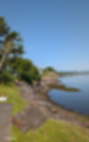 Rocky shoreline with trees on the left, calm blue water on the right, and a clear sky. A path and boulders line the edge.