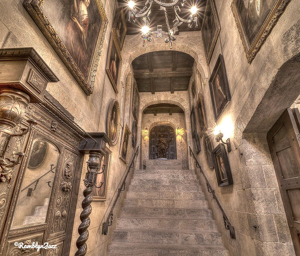Grand stone staircase with ornate framed portraits, an elegant chandelier above. Antique decor and warm lighting create a historic atmosphere.