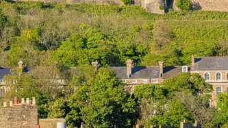 Castle on hill above historic town, red-cross flag flying, stone walls and green slopes under a clear blue sky.