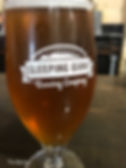 A glass of amber beer with "Sleeping Giant Brewing Company" logo, set on a wooden table with a blurred industrial background.