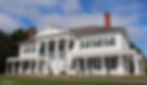 White colonial-style house with tall columns, green shutters, and red chimneys under a blue sky. Porch with chairs and neatly trimmed lawn.