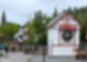 Clock tower reading Kimberley B.C. with red and white design, surrounded by trees and buildings. Cloudy sky, few people walking nearby.
