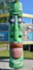 Green wooden totem with large lips and eyes, outdoors in front of informational signs. Text reads "Gitchi Goomee." Bright, sunny day.