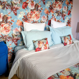 Floral bedroom with blue rose wallpaper, pastel pillows, striped white bedspread, and matching floral bedding;