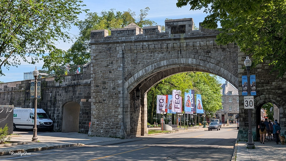 Stone archway with a road passing through. Festival banners and trees in the background. Streetlights and a speed limit sign are visible.