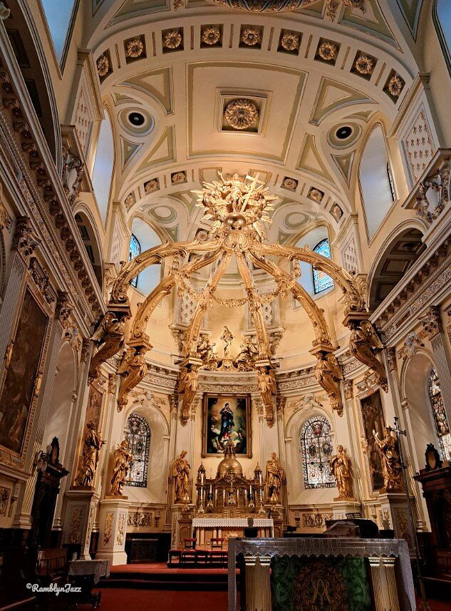 Ornate church interior with gold details, elaborate arches, and statues. Stained glass windows and an intricate ceiling adorn the setting.