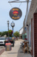 Street view with McLean's Vinyl Shop sign, vintage clock, and sidewalk sale sign. Shops and flowers line the street under a clear sky.