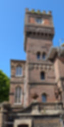 Ornate brick tower with arched windows and crenellations against a clear blue sky, beside a statue and stone balustrade.