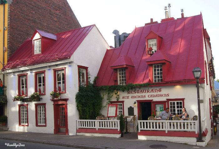 Charming red-roofed restaurant with white facade. "Restaurant Aux Anciens Canadiens" sign; diners on patio. Cozy, historic ambiance.