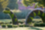 Topiary shaped like a green sea serpent with spikes in a park setting. Surrounded by shrubbery and trees, a building visible in the background.