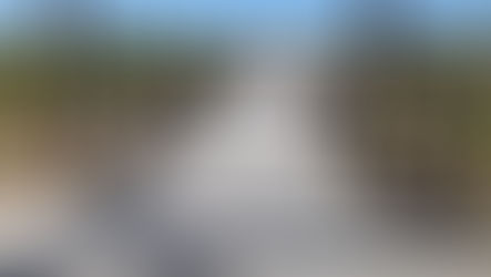 Wooden boardwalk under a slatted pergola leads to a sunny beach and blue ocean. Shadows create patterns on the path. Calm, inviting scene.