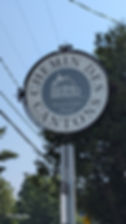 Round sign reading "Chemin des Cantons, Townships Trail" with a house illustration. Clear blue sky and trees in the background.