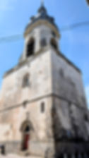 Tall weathered stone church tower with domed spire and red door against clear blue sky, crossed by overhead wires.