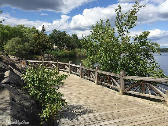 Wooden walkway by a river, surrounded by lush trees and rocks. Partly cloudy sky adds a serene, natural feel. No text visible.